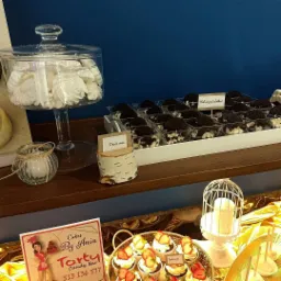 Słodki stół z deserami: bezy w szklanym naczyniu, ciasteczka oreo w plastikowych pojemnikach z napisem 'Miłość jest słodka!', babeczki z truskawkami na złotej tacy, świeca w ozdobnej klatce...