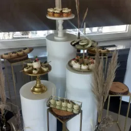 Elegancka ekspozycja deserów na białych cylindrach i złotych stojakach z drewnianymi elementami, minimalistyczna dekoracja z traw pampasowych w szklanych wazonach, czarno-biała podłoga.