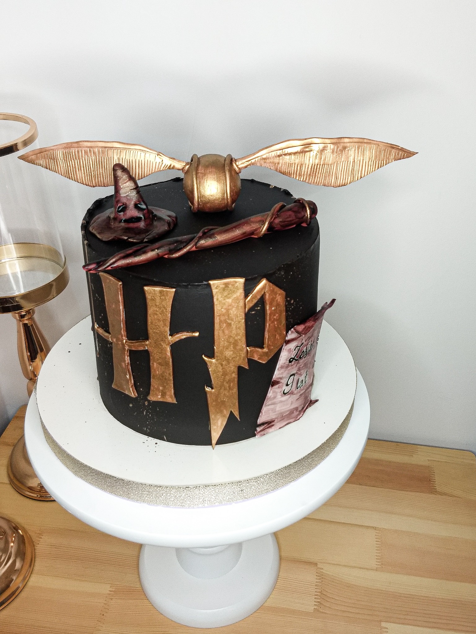 Czarny tort z motywami Harry'ego Pottera: Złoty Znicz, Tiara Przydziału, różdżka i zwój z napisem na białym stojaku.