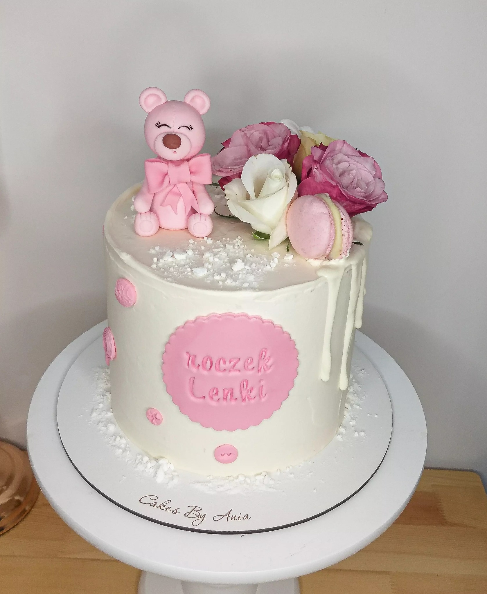 Tort urodzinowy dla dziewczynki z różowym misiem, kwiatami i makaronikiem, z napisem 'roczek Lenki' na różowym tle.