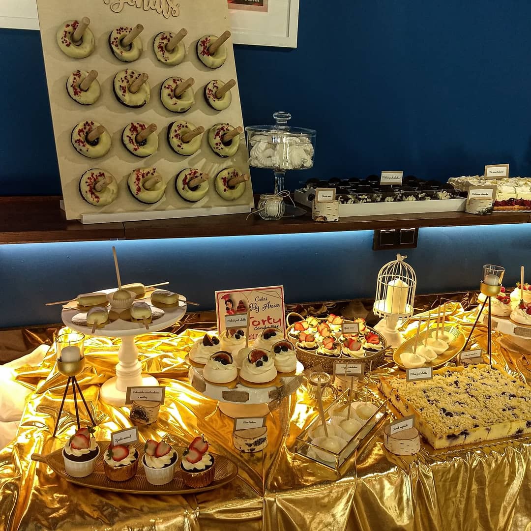 Stół z deserami na złotym obrusie: donuty na drewnianej ściance, babeczki z truskawkami, mini-torty z owocami, kostki z kremem, ciasto z kruszonką i jagodami, bezy w szklanym słoju, świece...