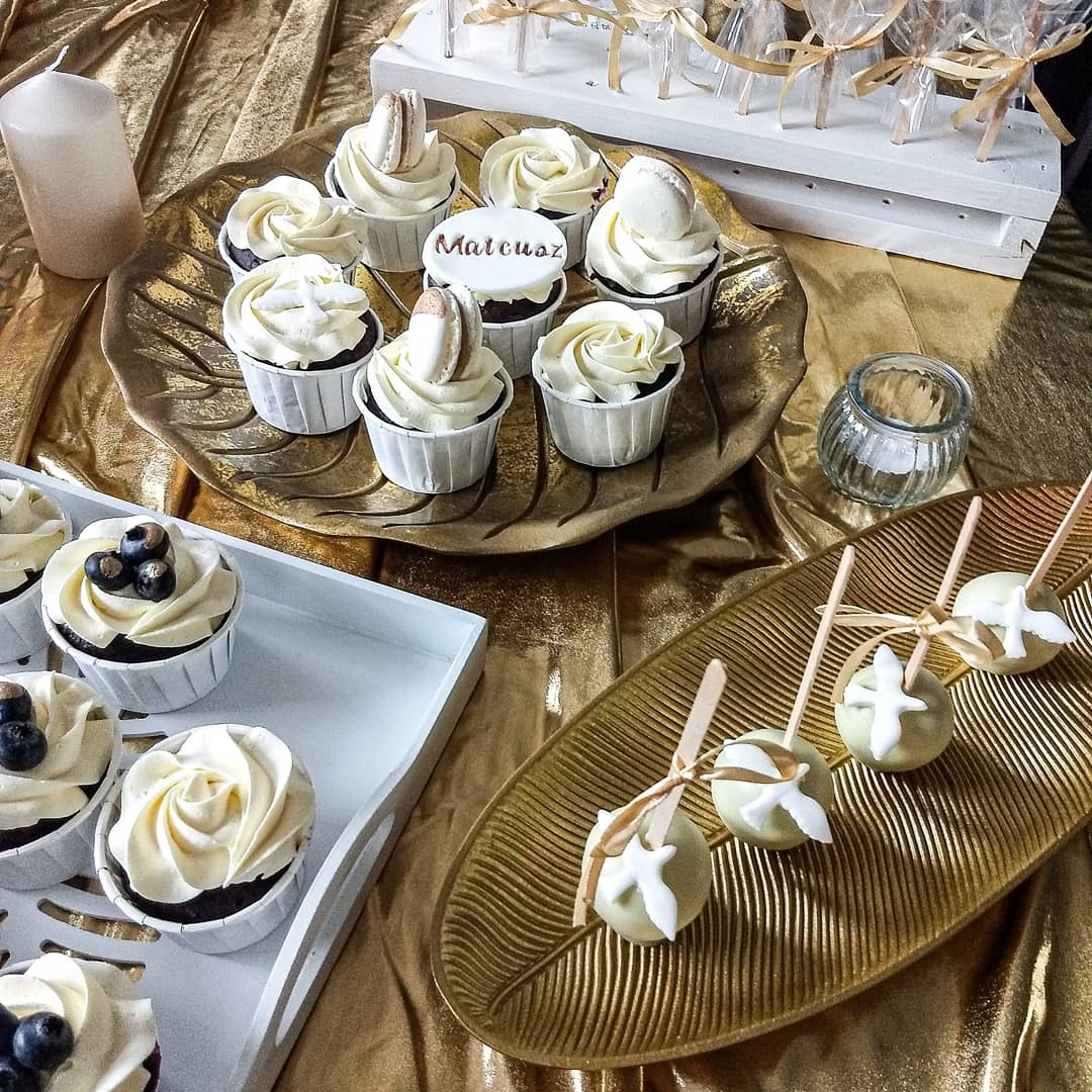 Dekoracyjne babeczki z kremem, makaronikami i napisem 'Mateusz' oraz cake pops na złotych paterach w kształcie liści, na tle błyszczącej, złotej tkaniny.