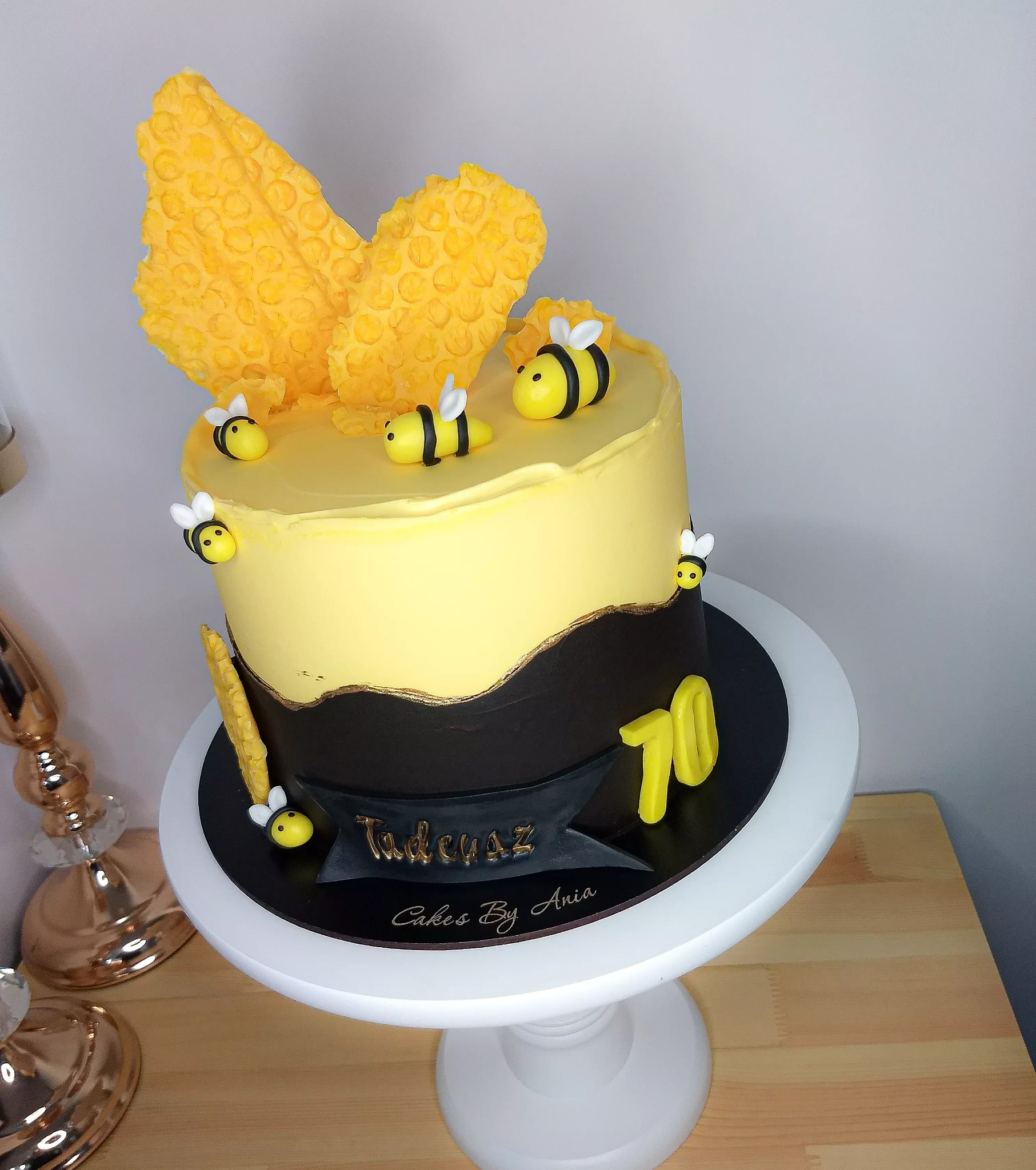 Tort urodzinowy z motywem pszczół, dwukolorowy krem (żółty i brązowy), dekoracje z wafli i lukru na białym stojaku.