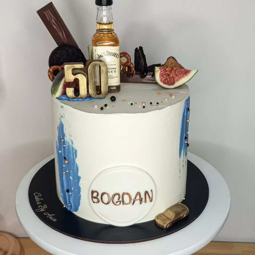 Tort urodzinowy w białym kremie z niebieskimi akcentami, dekorowany czekoladą, preclami, figą, butelką Jack Daniel's i złotym napisem '50' oraz imieniem 'BOGDAN'.