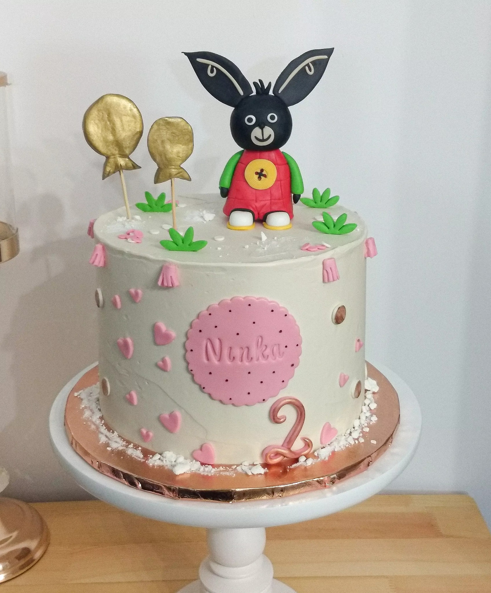 Tort urodzinowy z figurką Binga, złotymi balonami i imieniem 'Ninka' na różowym tle, dekorowany małymi różowymi serduszkami i zielonymi elementami, umieszczony na białym stojaku.