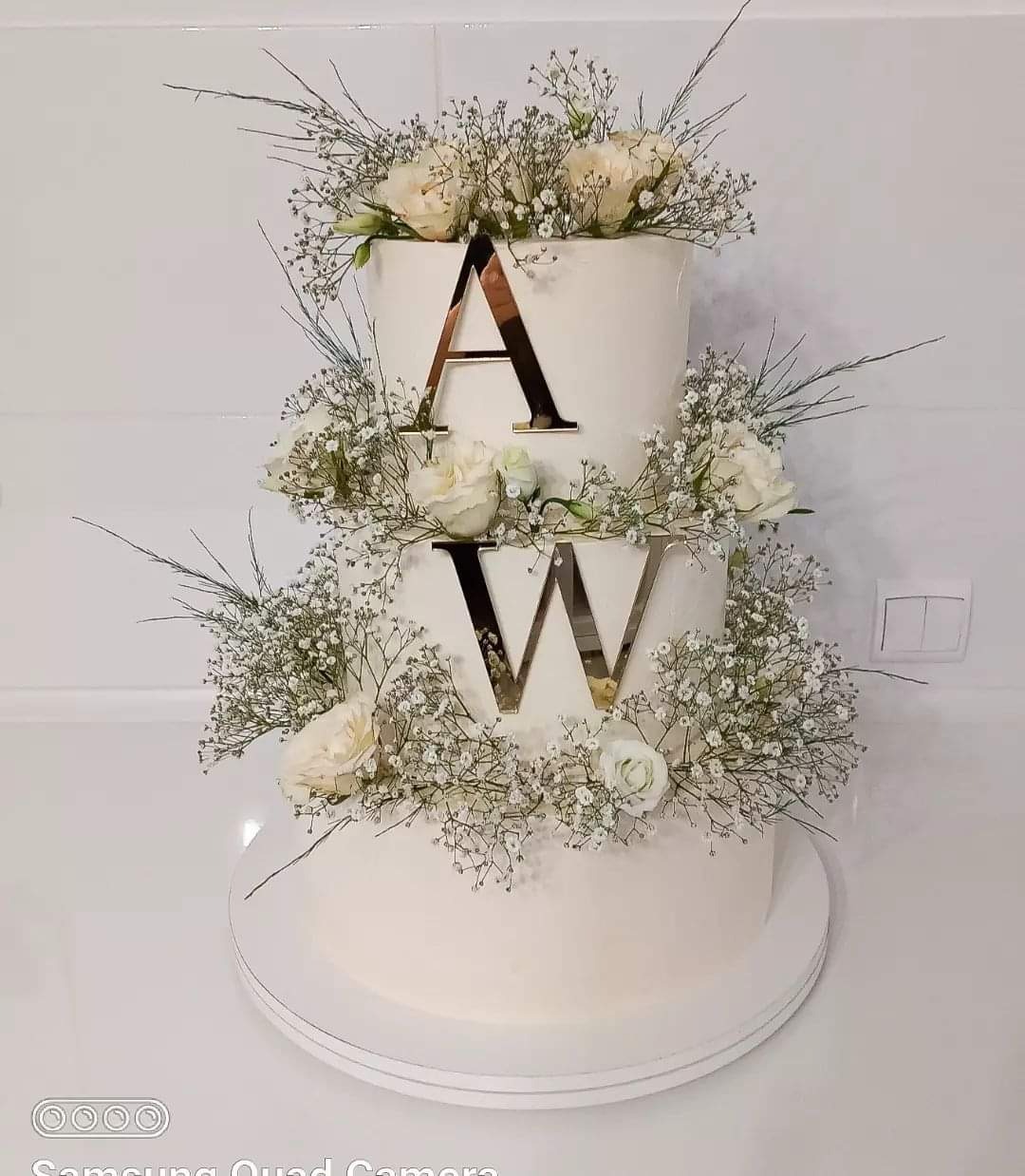 Dwupiętrowy tort weselny z białego kremu, ozdobiony białymi różami i gipsówką, z lustrzanymi literami 'A' i 'W' na każdym piętrze.