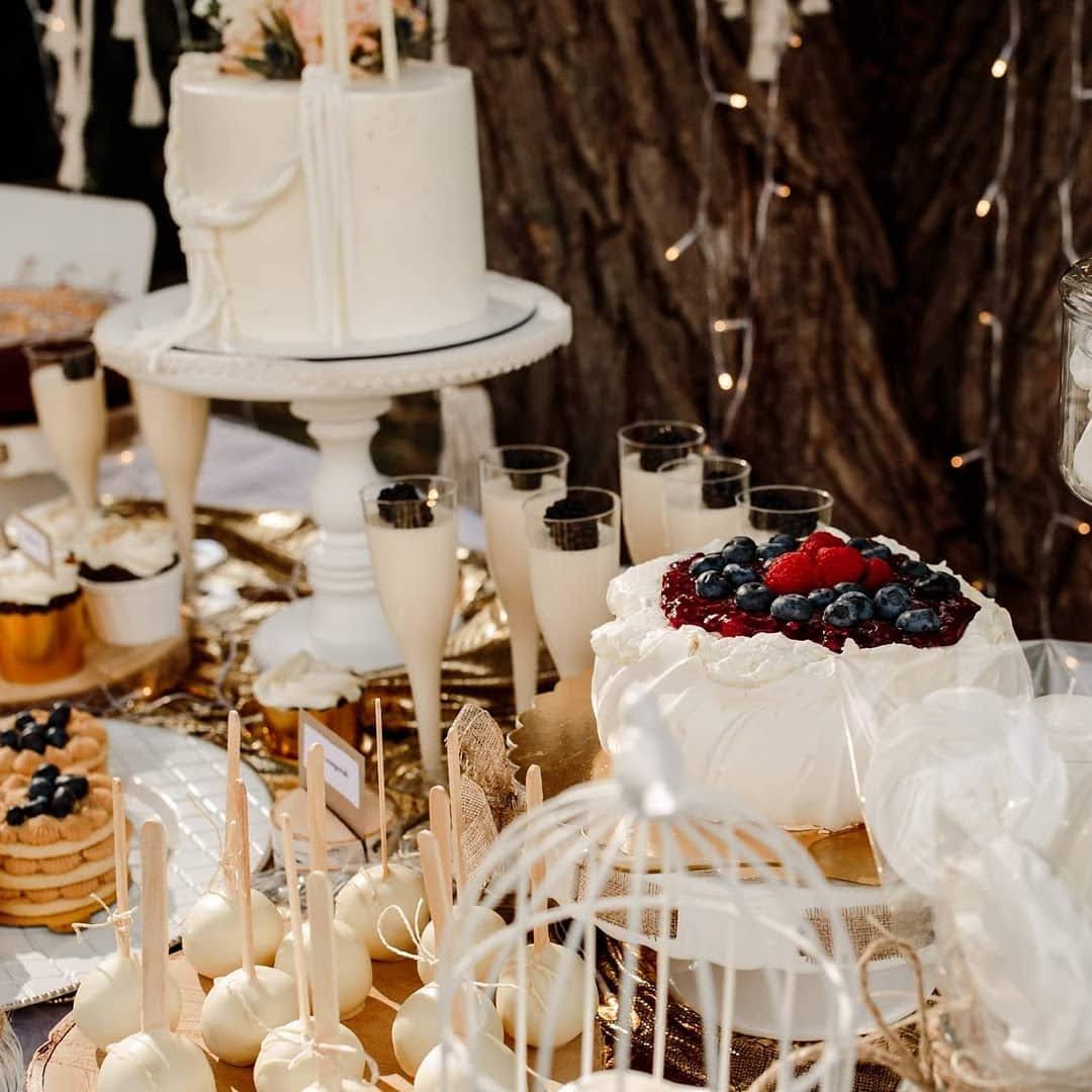 Stół z deserami na przyjęciu plenerowym: tort śmietanowy z owocami, babeczki w złotych papilotkach, cake pops w białej polewie, desery w kieliszkach, dekoracje w stylu rustykalnym i lampki.