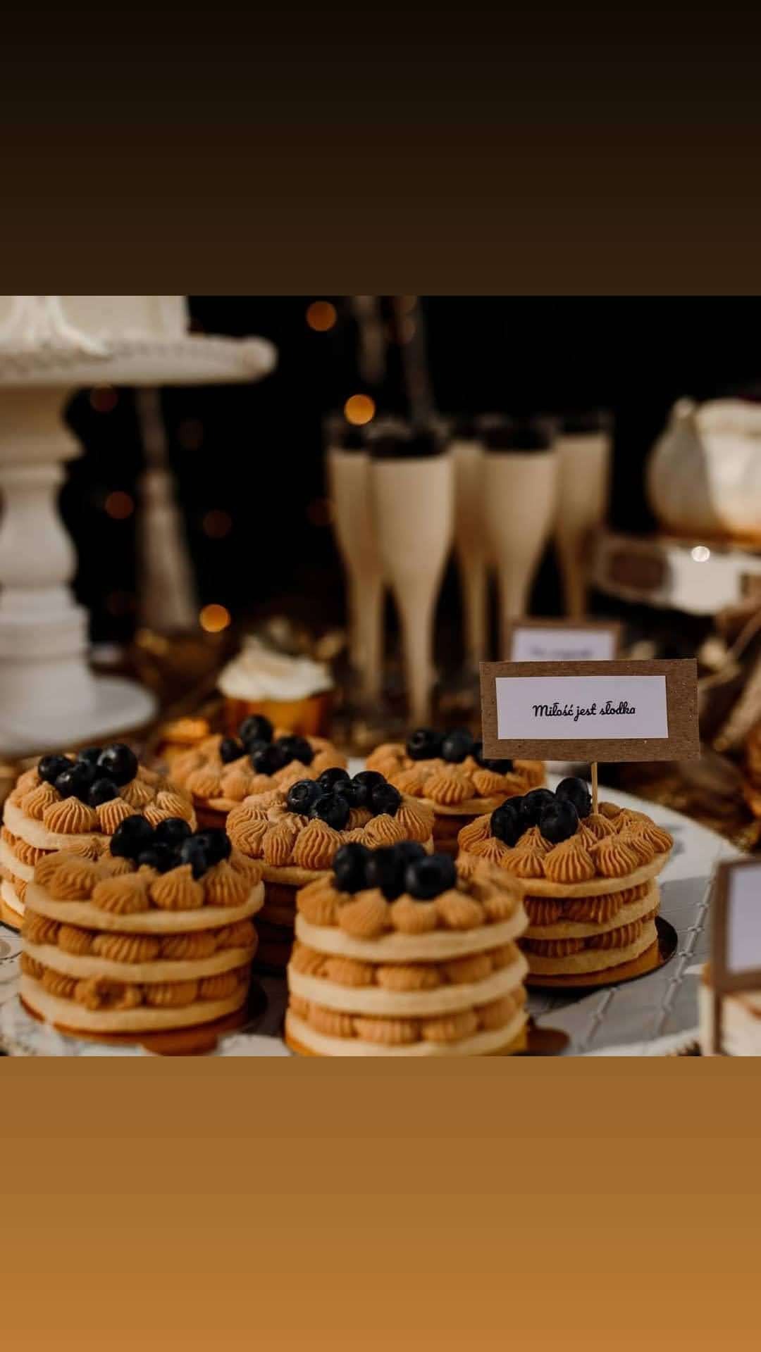 Miniaturowe torty z kremem i borówkami na stole deserowym, obok kieliszki z deserem i tabliczka z napisem 'Miłość jest słodka'.
