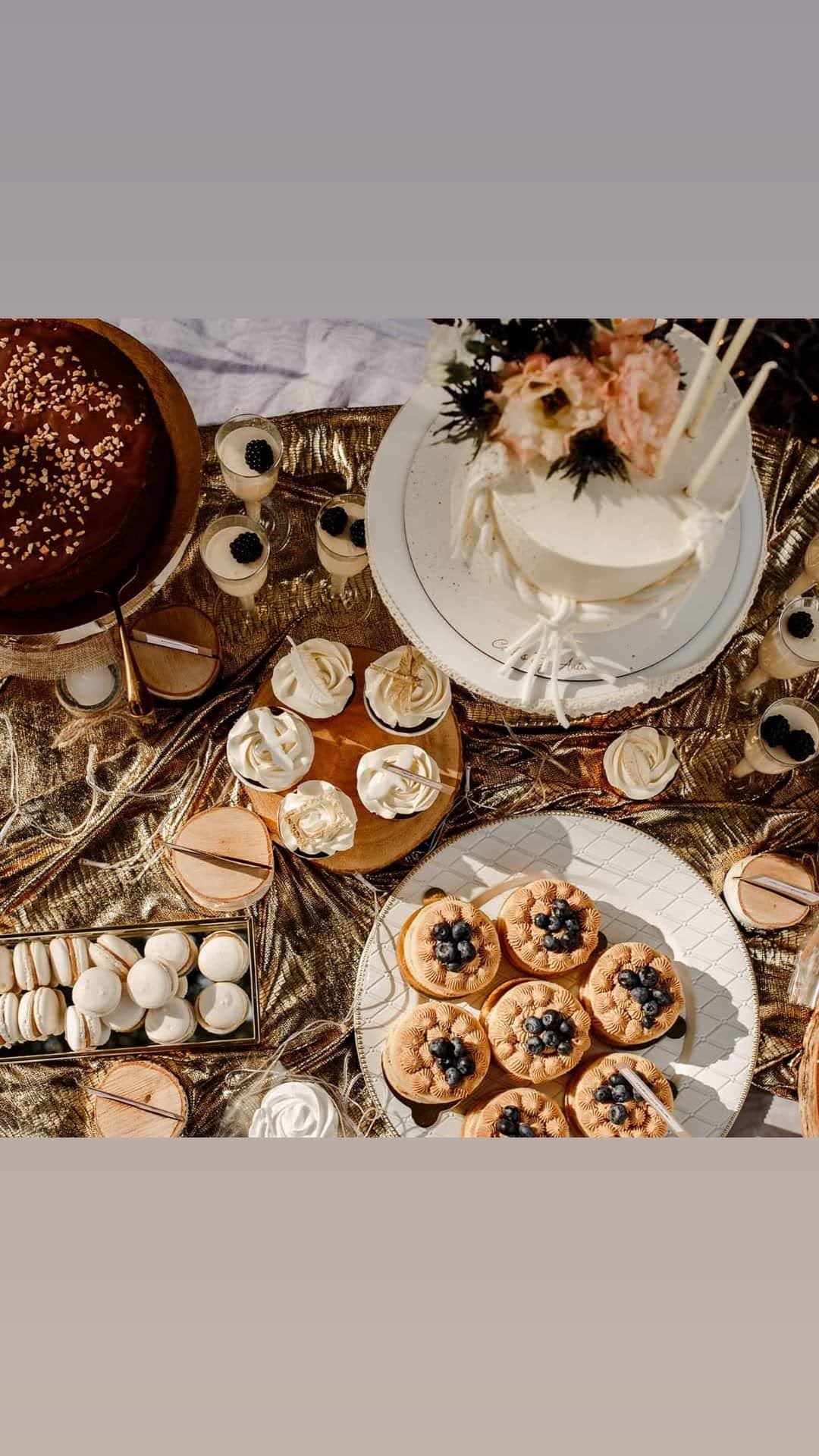 Ekskluzywny stół z deserami: tort z kwiatami, babeczki z kremem, makaroniki, desery w szklankach z jeżynami, wszystko na złotej, błyszczącej tkaninie.