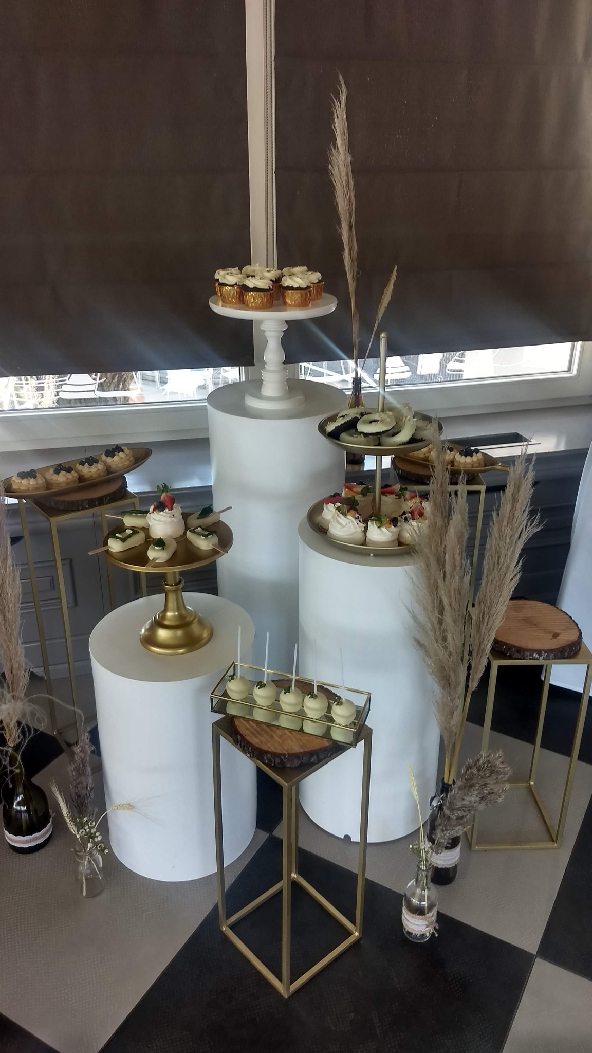 Elegancka ekspozycja deserów na białych cylindrach i złotych stojakach z drewnianymi elementami, minimalistyczna dekoracja z traw pampasowych w szklanych wazonach, czarno-biała podłoga.