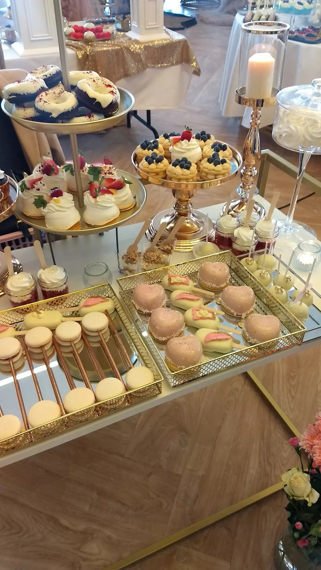 Elegancki stół z różnorodnymi deserami: donuty z białą polewą, bezy z truskawkami, babeczki z borówkami, mini desery w słoiczkach, ciastka na patyku w kształcie serca, makaroniki na patyku, lizaki.