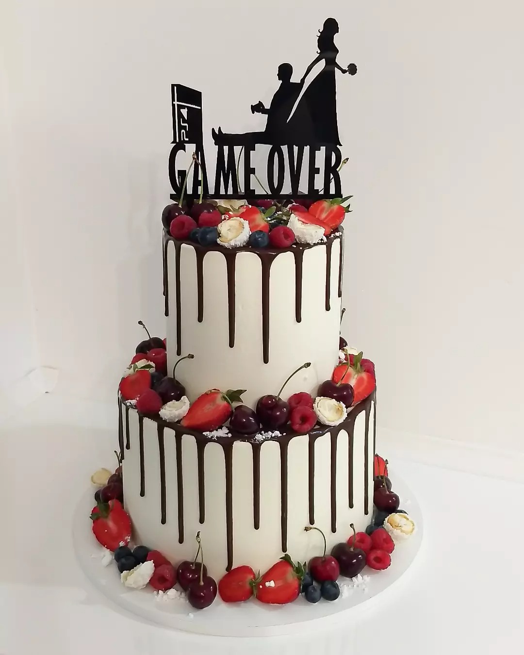 Dwupiętrowy tort weselny z białym kremem, polewą czekoladową, dekoracją z truskawek, wiśni, malin i borówek, zwieńczony czarnym topperem z napisem 'GAME OVER' i sylwetkami pary młodej oraz konsoli...