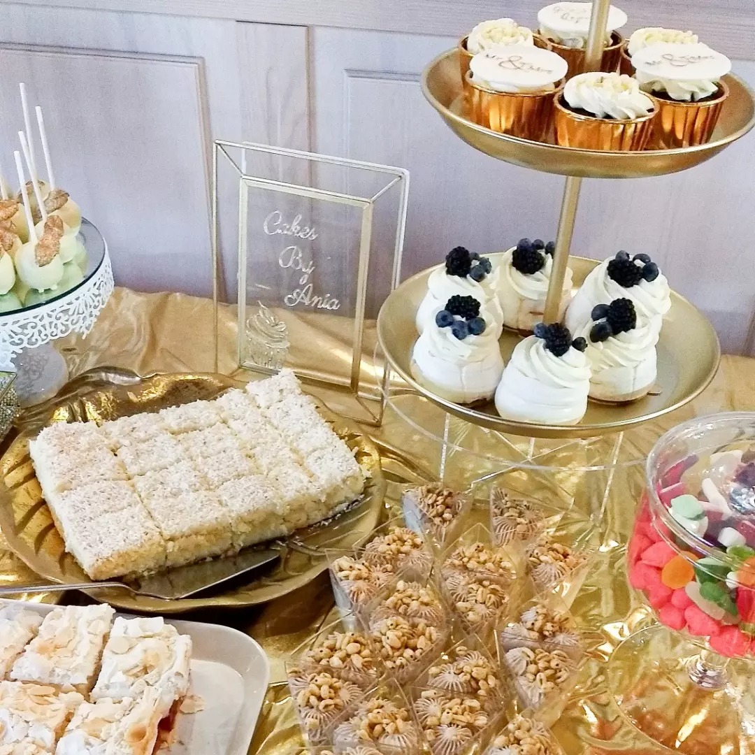 Słodki stół z różnorodnymi deserami: mini bezy z owocami, babeczki, ciasto kokosowe w kwadratach, desery w małych pojemnikach z orzechami i kolorowe żelki, złota dekoracja stołu i ramka z napisem...