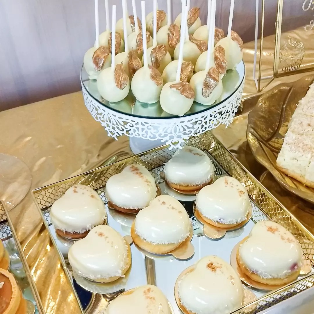 Dekoracyjny stół z deserami: lizaki w białej czekoladzie ze złotymi akcentami na ażurowym stojaku, obok ciasteczka w kształcie serc z białą polewą i złotymi drobinkami, całość w złotej tonacji.