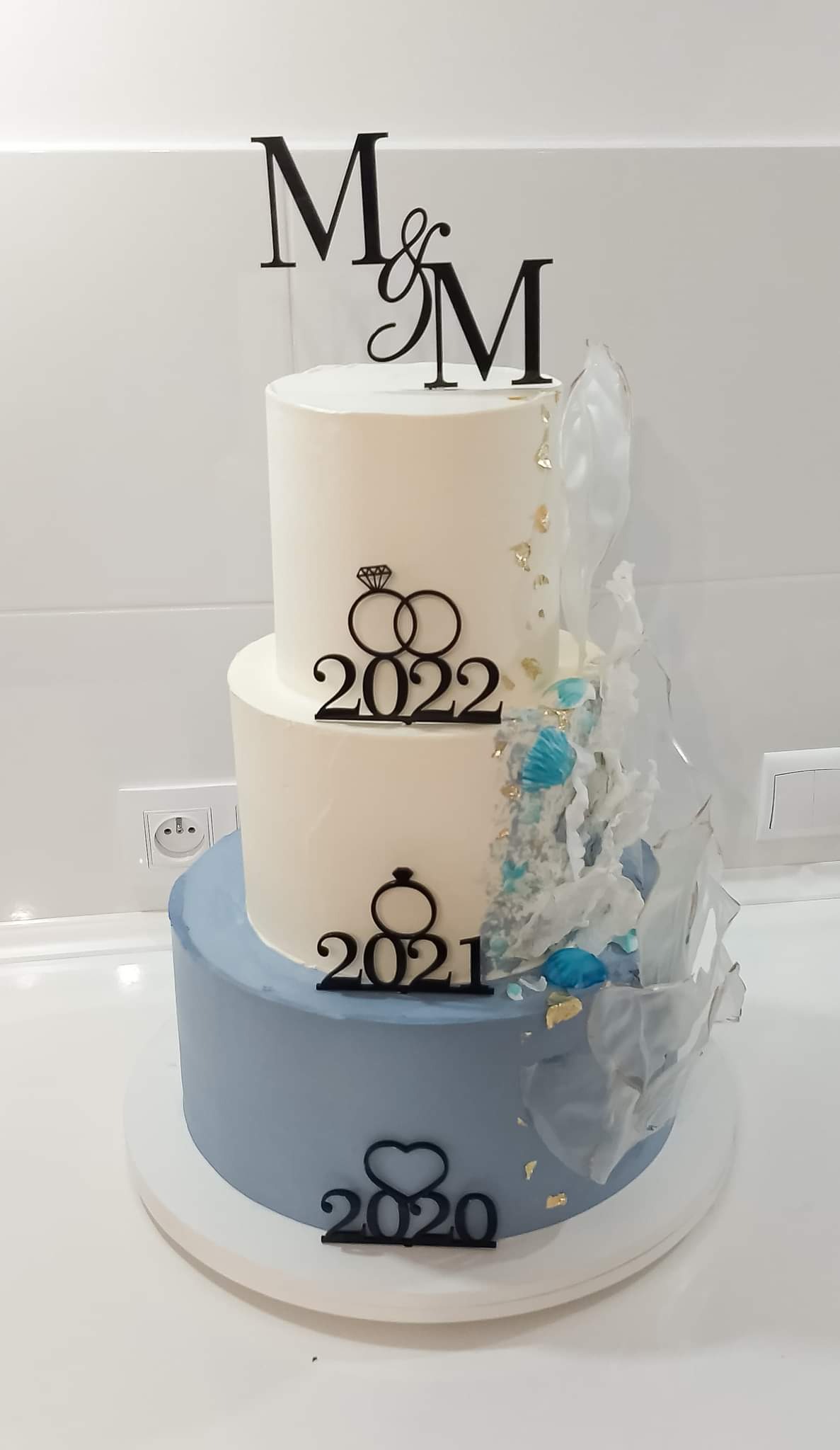 Trzypiętrowy tort weselny w odcieniach bieli i błękitu, ozdobiony jadalnymi dekoracjami przypominającymi szkło, złotymi płatkami, topperem z inicjałami 'M&M' oraz datami 2020, 2021 i 2022...