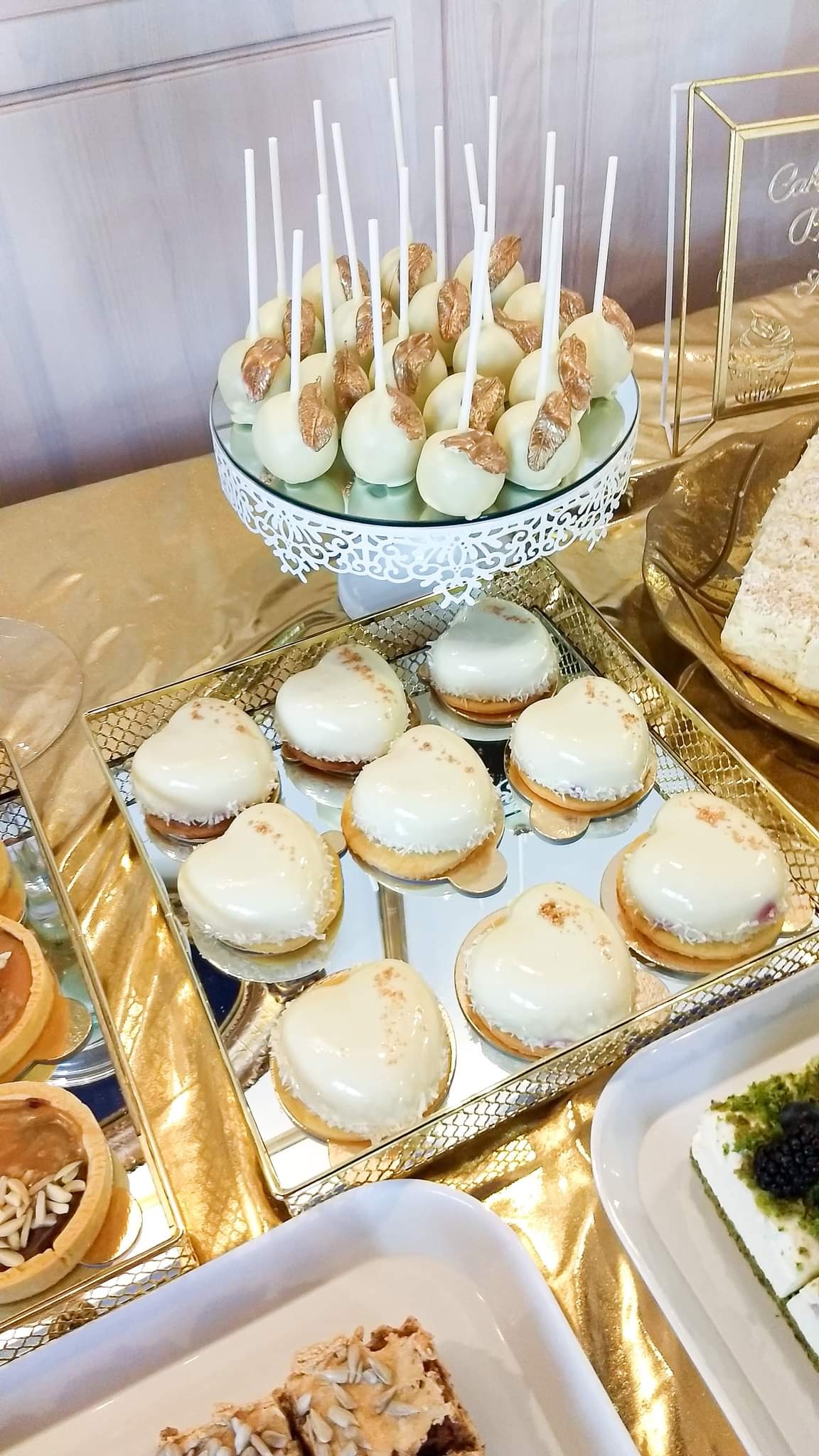 Elegancki bufet ze słodkościami: cake pops z białej czekolady i złotymi zdobieniami na ażurowym stojaku, serca z białej glazury na złotej tacy, tartaletki i ciasta na złotym obrusie.