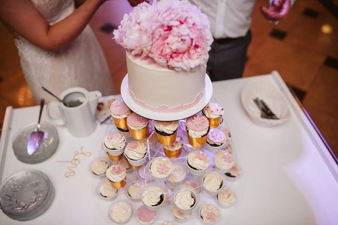 Dwupoziomowy tort ślubny z białego kremu z dekoracją z różowych piwonii na szczycie, otoczony babeczkami z kremem i różowymi kwiatkami, ustawiony na białym stole obok srebrnych naczyń i napisu 'Love'.