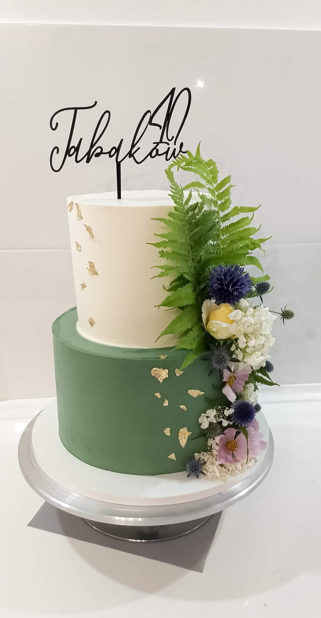 Dwupoziomowy tort w odcieniach zieleni i kremu, zdobiony złotymi płatkami, paprocią i kwiatami, z czarnym topperem z napisem 'Fabianów 40'.