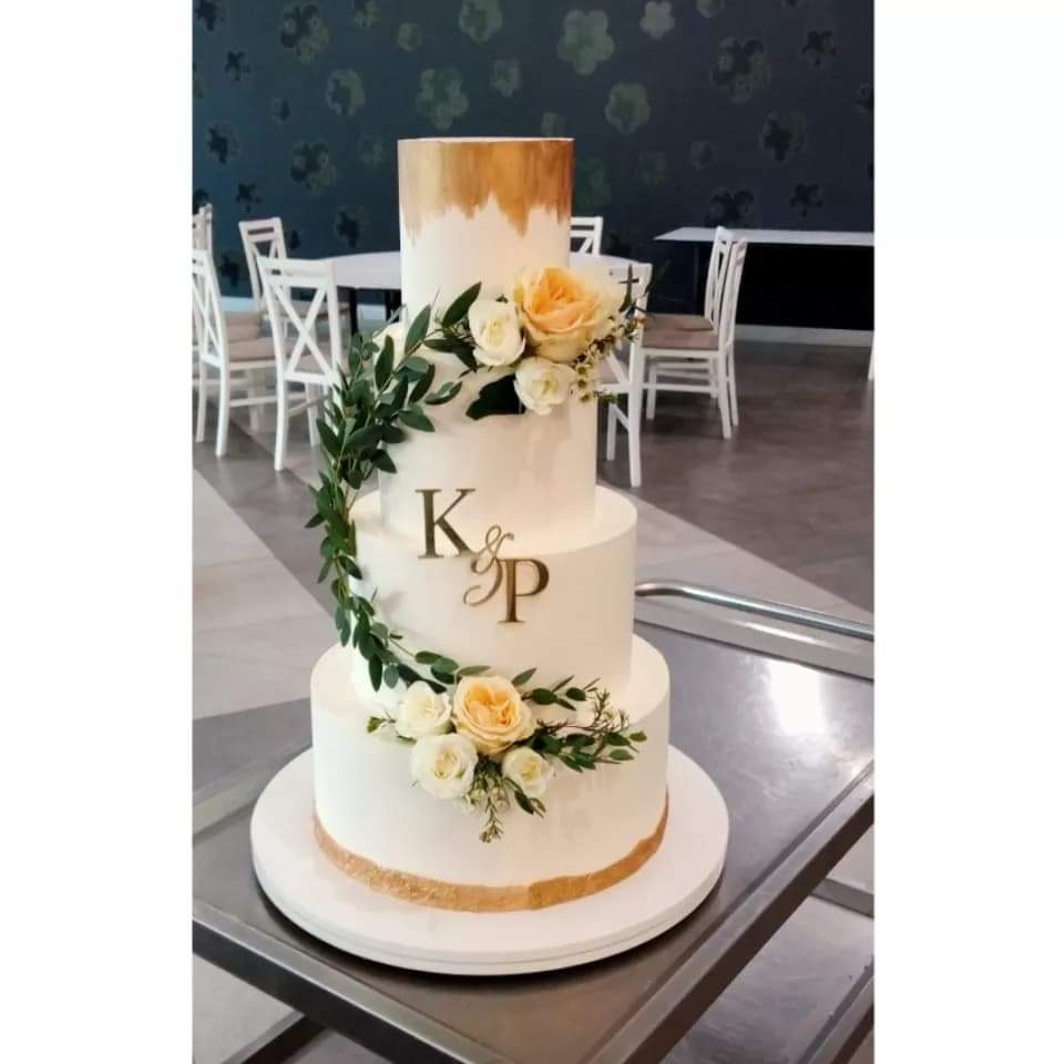 Trzypiętrowy tort weselny w białej polewie, zdobiony żywymi kwiatami w odcieniach bieli i kremu oraz złotymi akcentami, inicjały 'K&P' na środkowym piętrze, tło stanowią krzesła i stoliki.