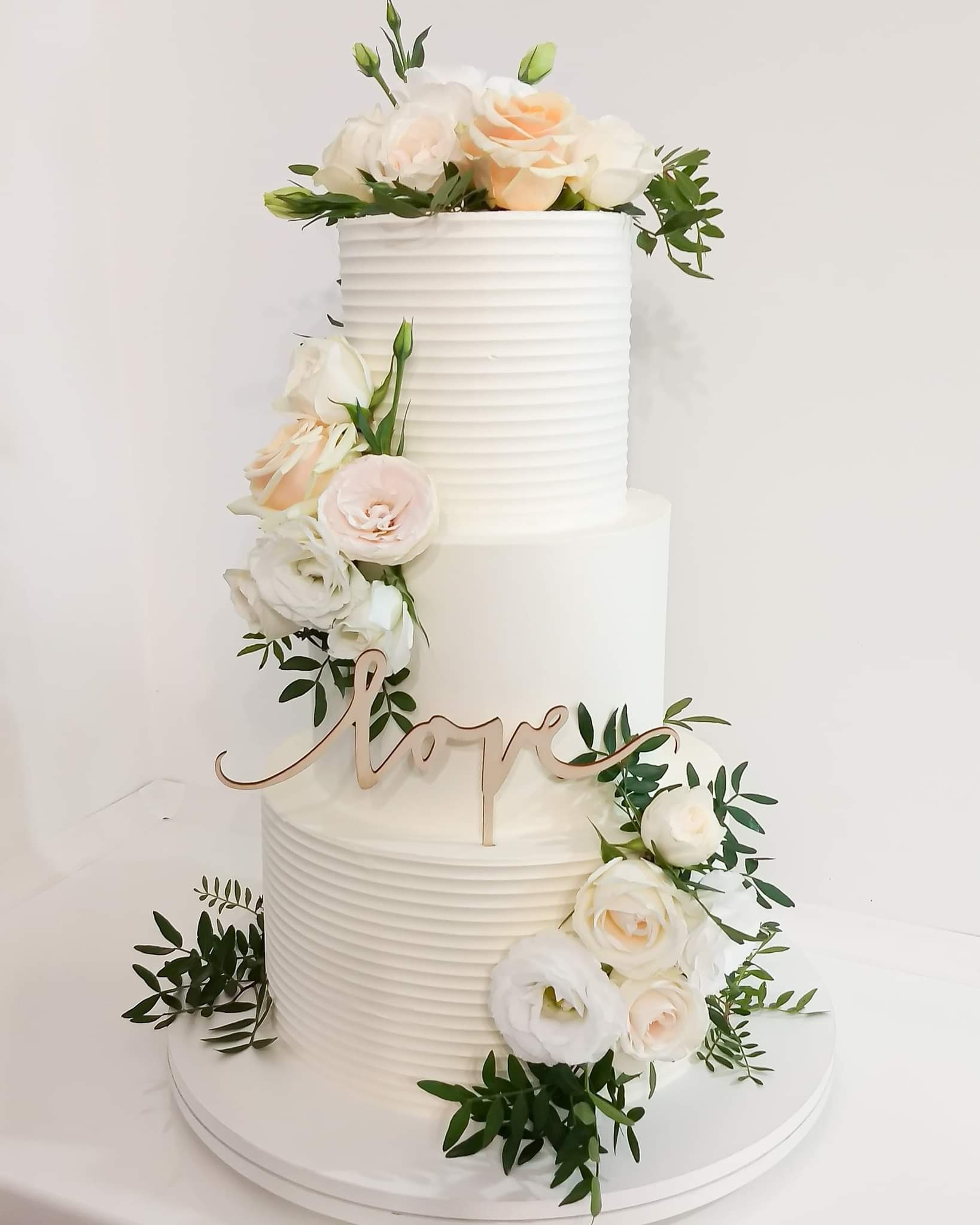 Trzypiętrowy tort weselny z białym kremem, zdobiony świeżymi, białymi i brzoskwiniowymi różami oraz drewnianym napisem 'love'.