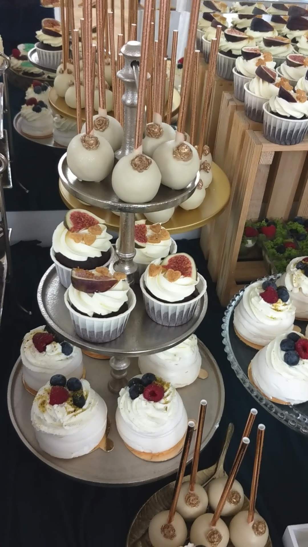 Elegancki, trzypoziomowy pater z deserami: babeczki z figami i kremem, mini bezy z owocami oraz cake pops z dekoracjami w kolorze złota i miedzianymi patyczkami.
