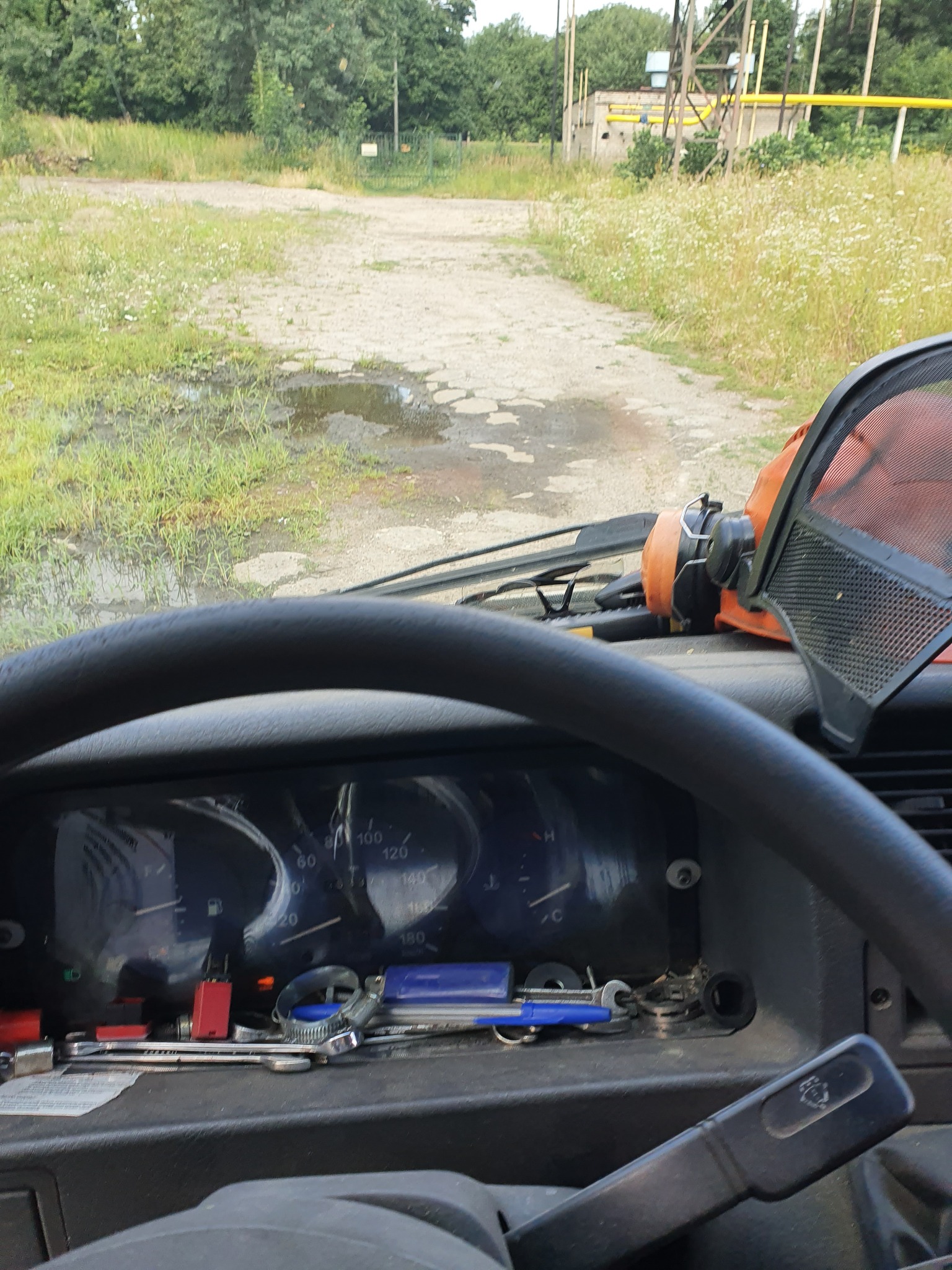 Widok z kabiny pojazdu na drogę prowadzącą obok zarośniętego terenu z widoczną infrastrukturą techniczną w tle, na desce rozdzielczej leżą narzędzia i osłona twarzy.