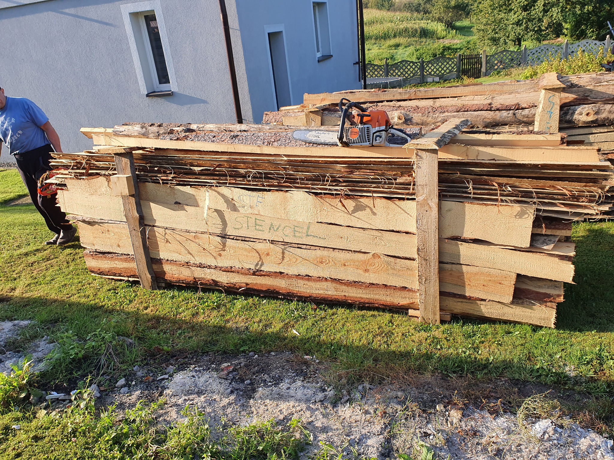 Stos desek z napisem 'STENCEL' spięty drewnianymi belkami, na wierzchu spalinowa piła łańcuchowa, w tle budynek i zieleń