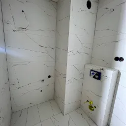 Świeżo wykafelkowana łazienka z białymi płytkami imitującymi marmur, widoczne otwory na instalacje i zabudowany stelaż podtynkowy WC z żółtymi rurami.