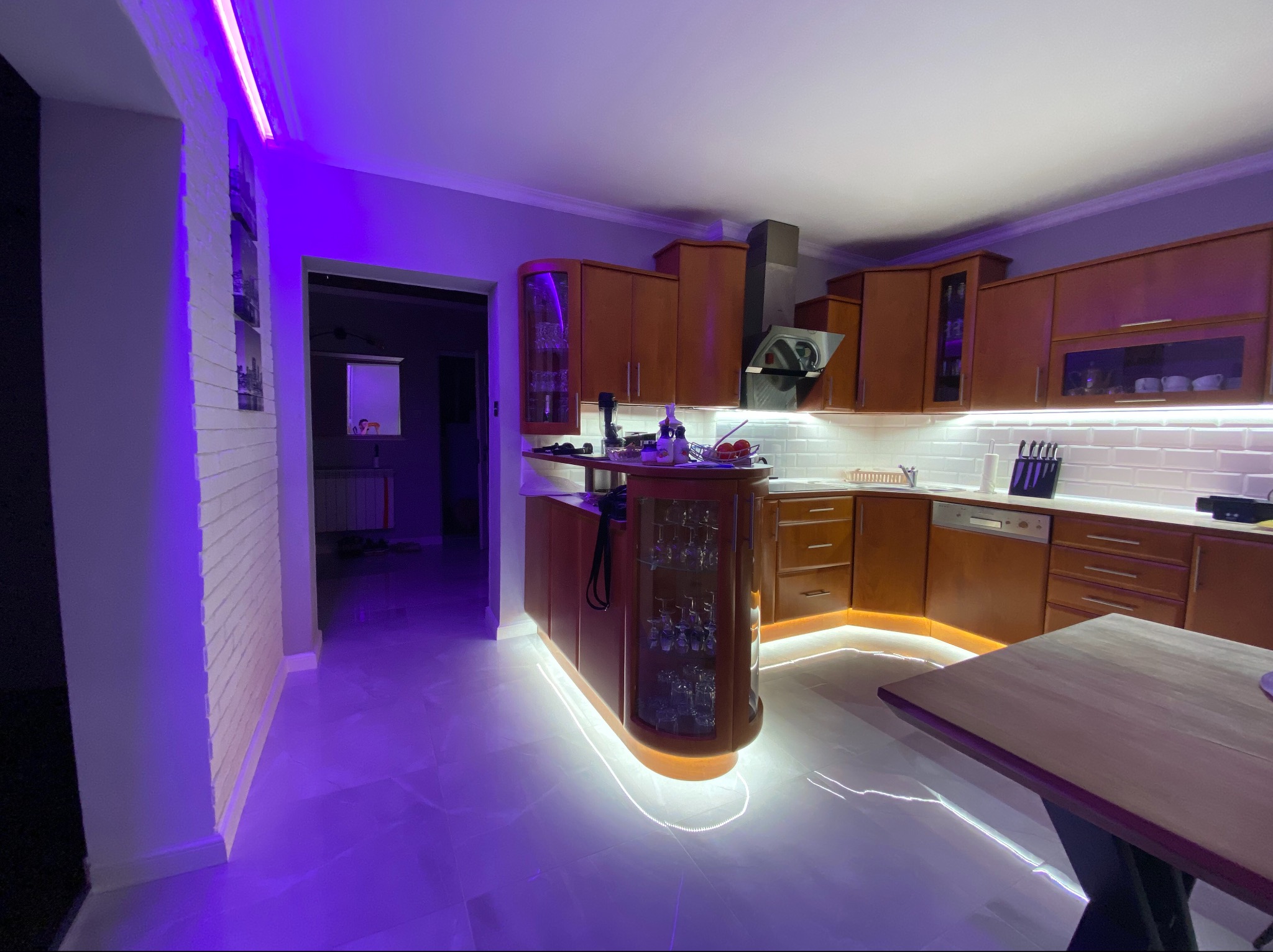 Nowoczesna kuchnia z drewnianymi szafkami, białymi płytkami na ścianie i podświetleniem LED w kolorze pomarańczowym pod szafkami i fioletowym na suficie, widoczny stół i przeszklona witryna...