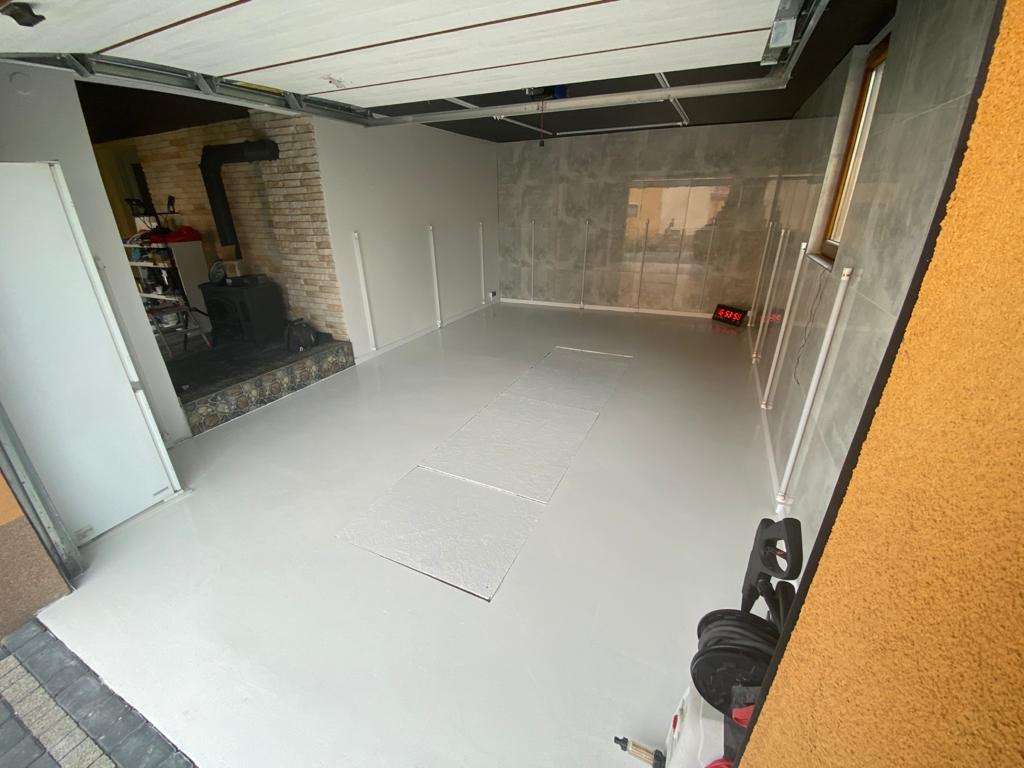 Wnętrze garażu po wylaniu jasnoszarej posadzki żywicznej, widoczny piec na drewno z ceglaną obudową w głębi pomieszczenia i cyfrowy zegar na ścianie.