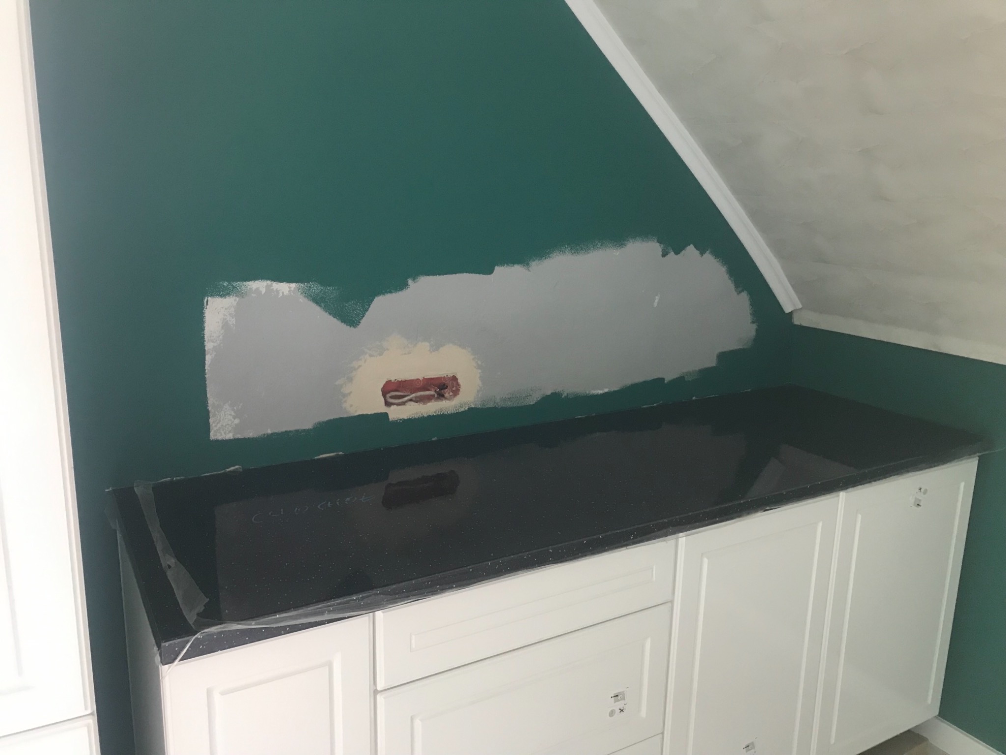 Montaż czarnego blatu z brokatem na białych szafkach kuchennych, ściana pomalowana na zielono z widocznym miejscem po instalacji elektrycznej w trakcie przygotowania do prac wykończeniowych.