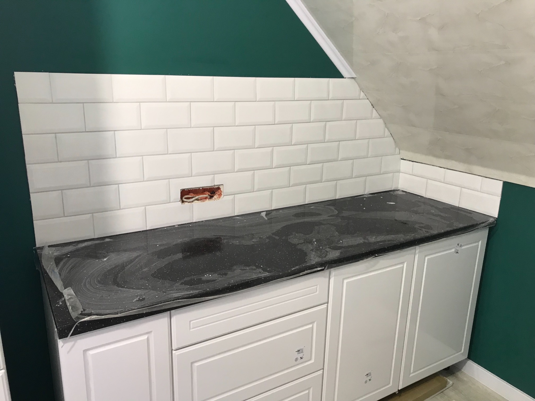 Częściowo wykończona kuchnia z białymi, prostokątnymi płytkami na ścianie, czarnym blatem roboczym z folią ochronną i białymi szafkami; widoczne gniazdko elektryczne.