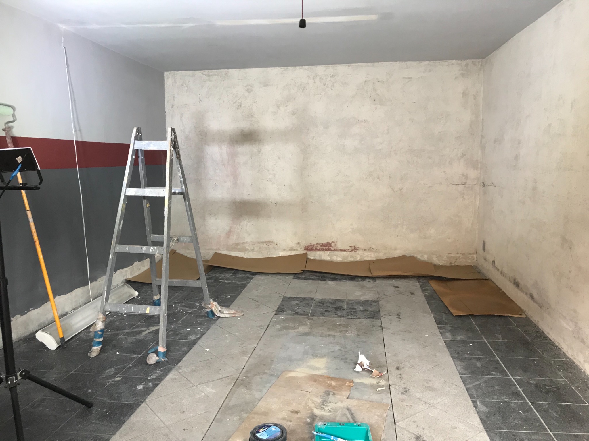 Pomieszczenie w trakcie remontu: drabina, zabezpieczona podłoga kartonem, ściany częściowo pomalowane, widoczne narzędzia malarskie.