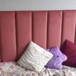 Tapicerowany panel zagłówka w odcieniach różu z pionowymi przeszyciami, widoczne poduszki dekoracyjne: biała z falbanami, fioletowa w kształcie gwiazdy z uśmiechem i bordowa, na łóżku z pościelą...