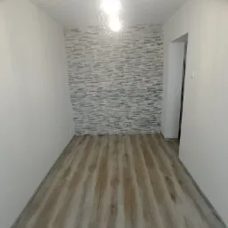 Wnętrze wykończonego pokoju z drewnopodobną podłogą, jedną ścianą wyłożoną dekoracyjną szarą cegłą i nowoczesną lampą wiszącą z odsłoniętą żarówką.