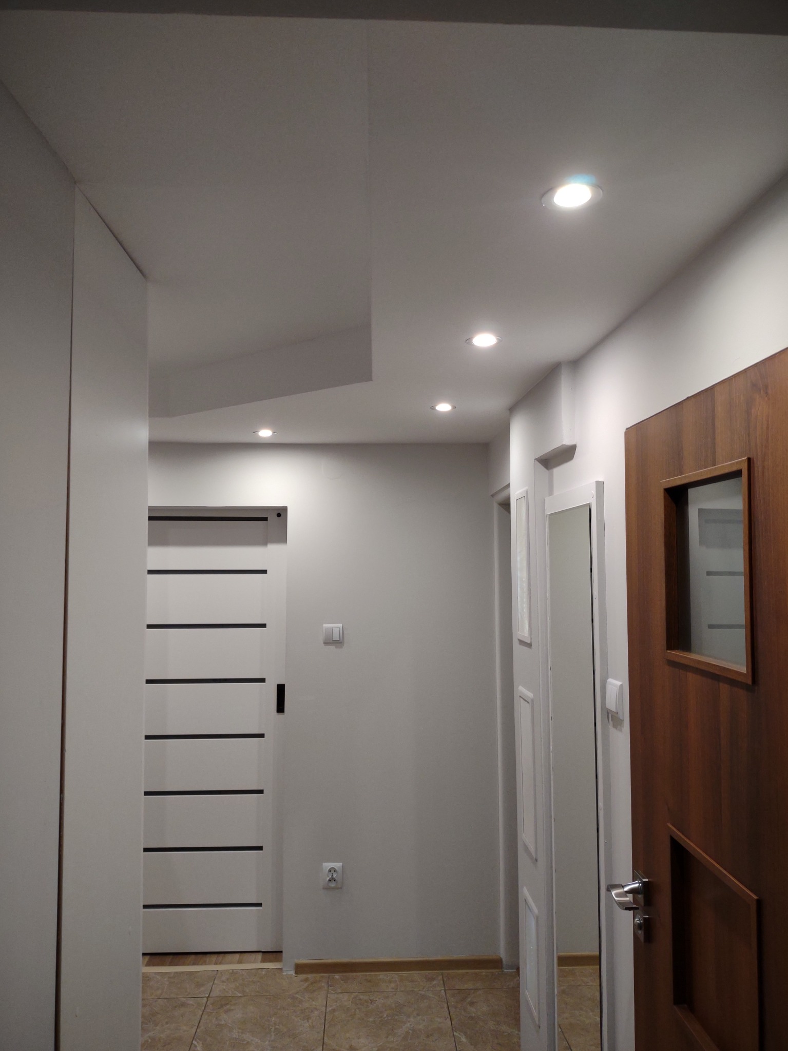 Nowoczesny korytarz z białymi ścianami, podwieszanym sufitem z oświetleniem punktowym, dwiema parami drzwi wewnętrznych - białe z czarnymi paskami i brązowe z przeszkleniem, oraz ceramiczną podłogą...