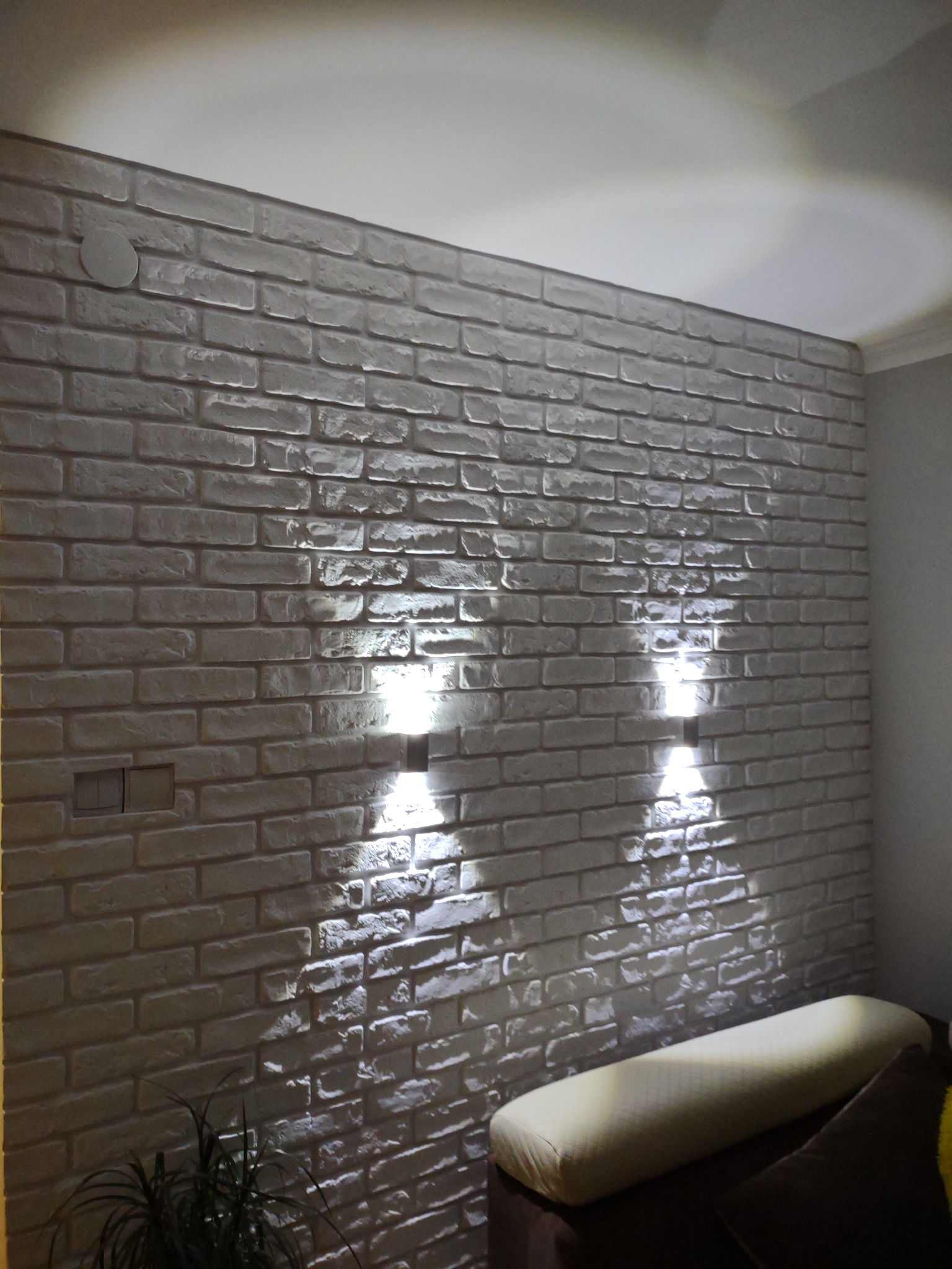 Ściana z dekoracyjnej białej cegły oświetlona przez dwa kinkiety, widoczne gniazdka elektryczne i fragment sofy z poduszką.
