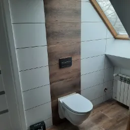Nowoczesna łazienka na poddaszu z drewnopodobnymi płytkami na podłodze i ścianie, wisząca toaleta i grzejnik pod oknem dachowym.