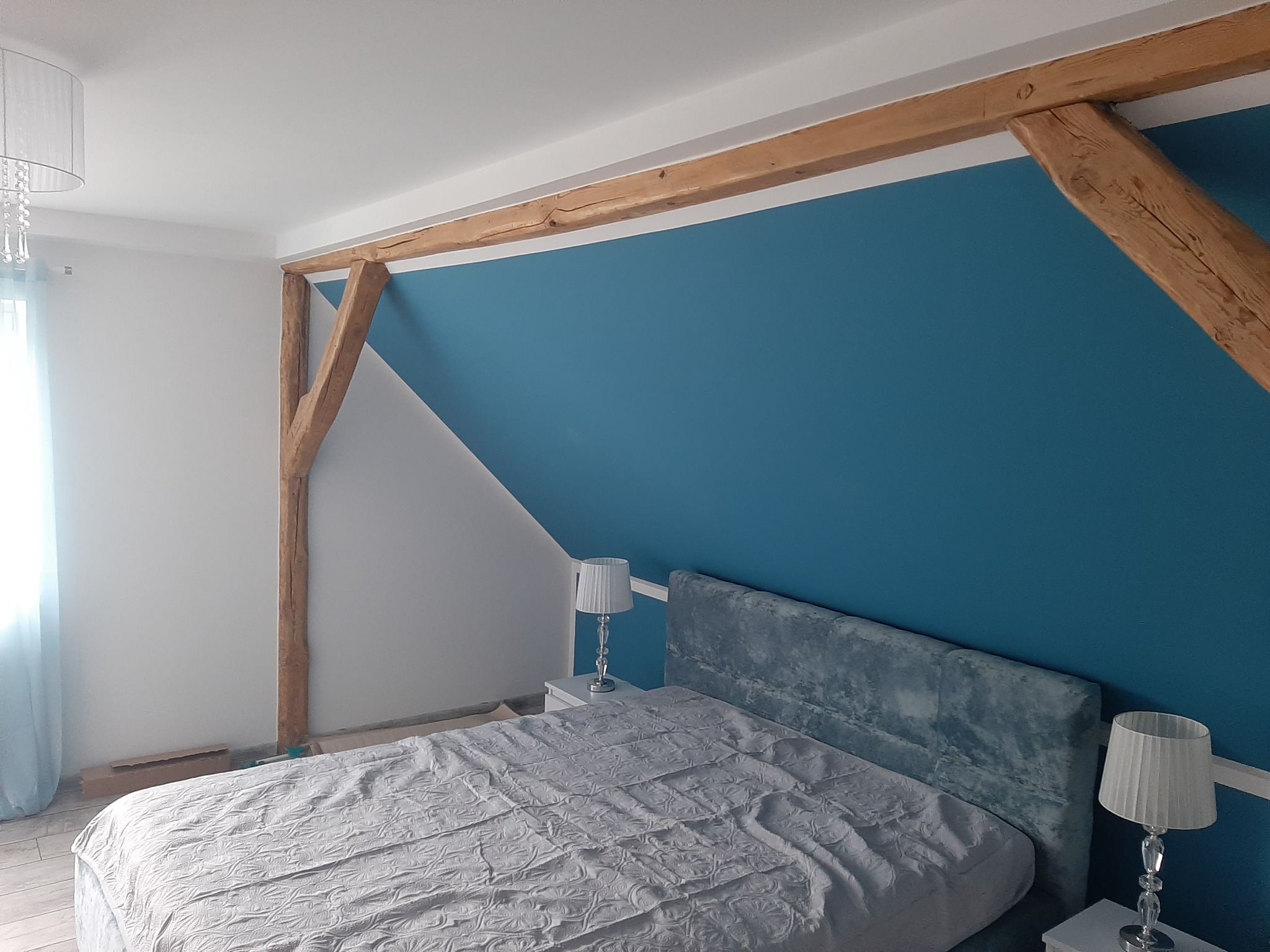Sypialnia na poddaszu z drewnianą konstrukcją stropu, pomalowaną na niebiesko ścianą i łóżkiem z szarym zagłówkiem.