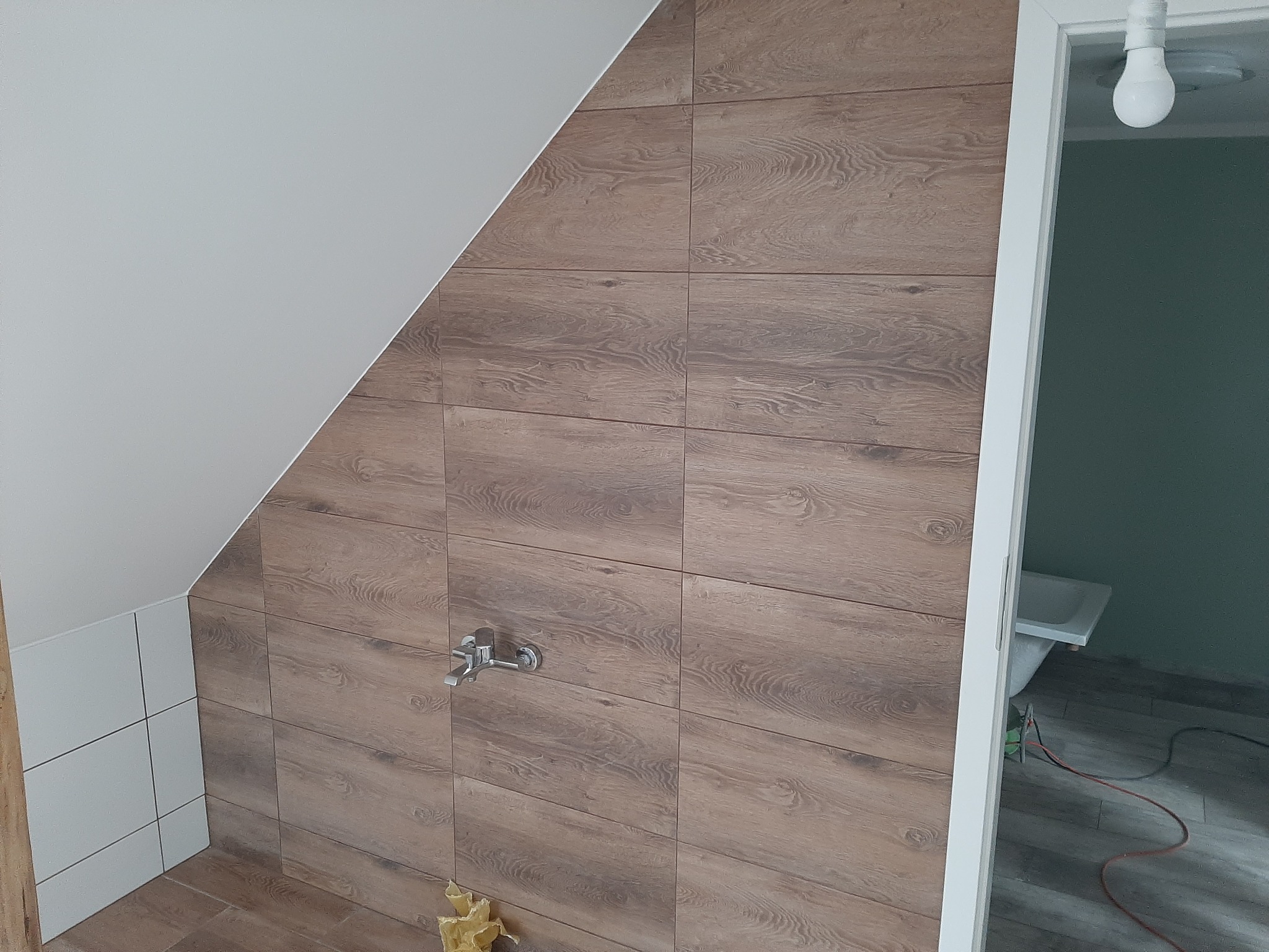 Wykończona ściana w łazience na poddaszu w miejscowości Wieleń, wyłożona brązowymi płytkami imitującymi drewno, z zamontowaną baterią. Widoczny fragment białej ściany i drzwi do łazienki.