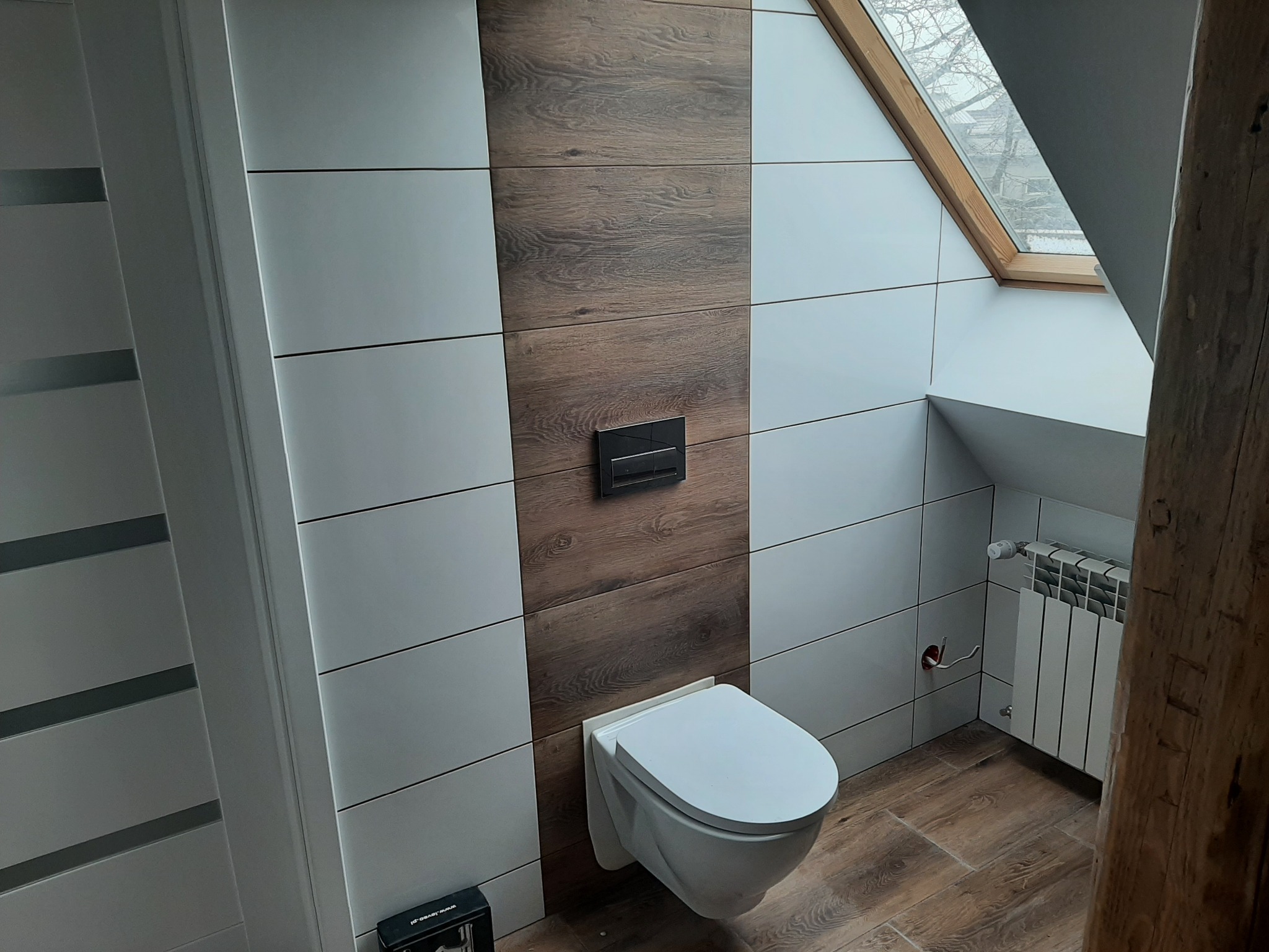 Nowoczesna łazienka na poddaszu z drewnopodobnymi płytkami na podłodze i ścianie, wisząca toaleta i grzejnik pod oknem dachowym.