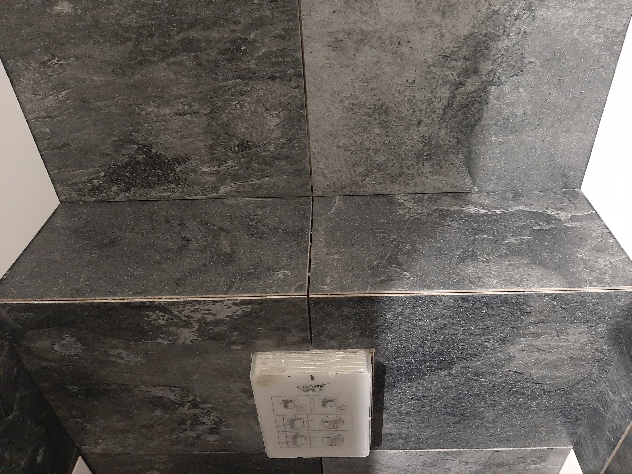 Szara, kamieniopodobna okładzina ceramiczna w łazience, z widocznym przyciskiem spłuczki WC marki Grohe, zamontowanym na ścianie.