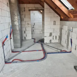 Instalacja hydrauliczna w surowym stanie, rury w izolacji czerwonej i niebieskiej, szare rury kanalizacyjne, ściany z białego gazobetonu, drewniana konstrukcja dachu z oknem dachowym, widok...