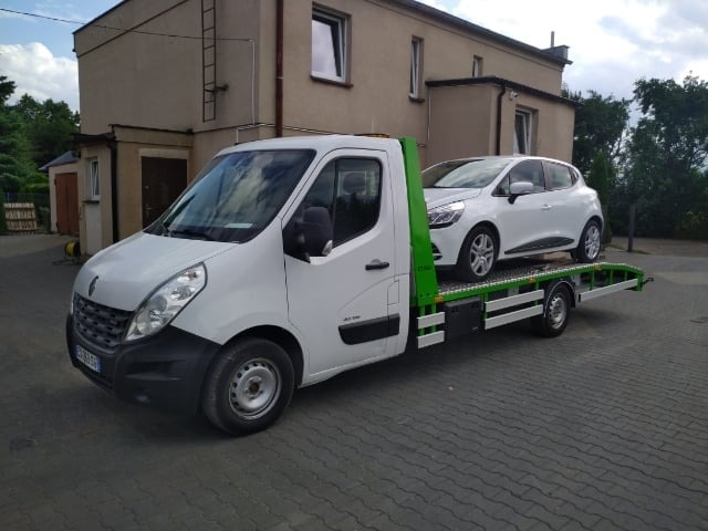 Biała laweta Renault Master z zielonym najazdem przewożąca biały samochód osobowy Renault Clio na tle budynku z beżową elewacją i brukowanej nawierzchni.