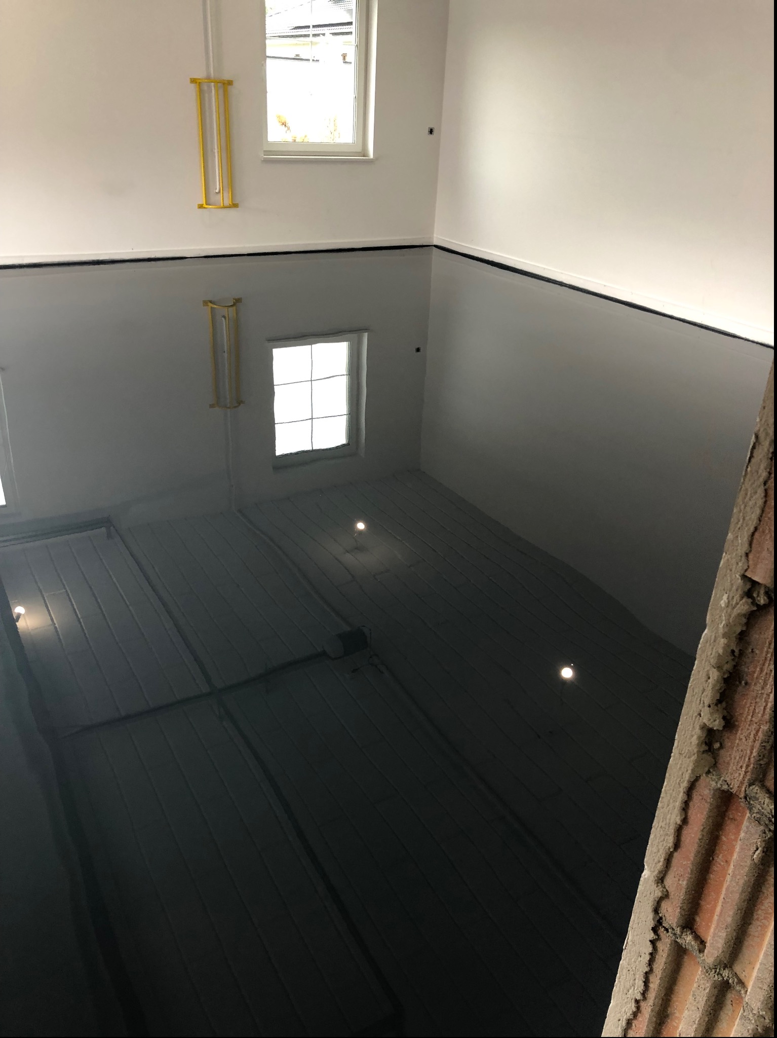 Błyszcząca, ciemna posadzka w jasnym pomieszczeniu z dwoma oknami i żółtymi elementami instalacji na ścianie. Widok z góry, fragment muru po prawej.