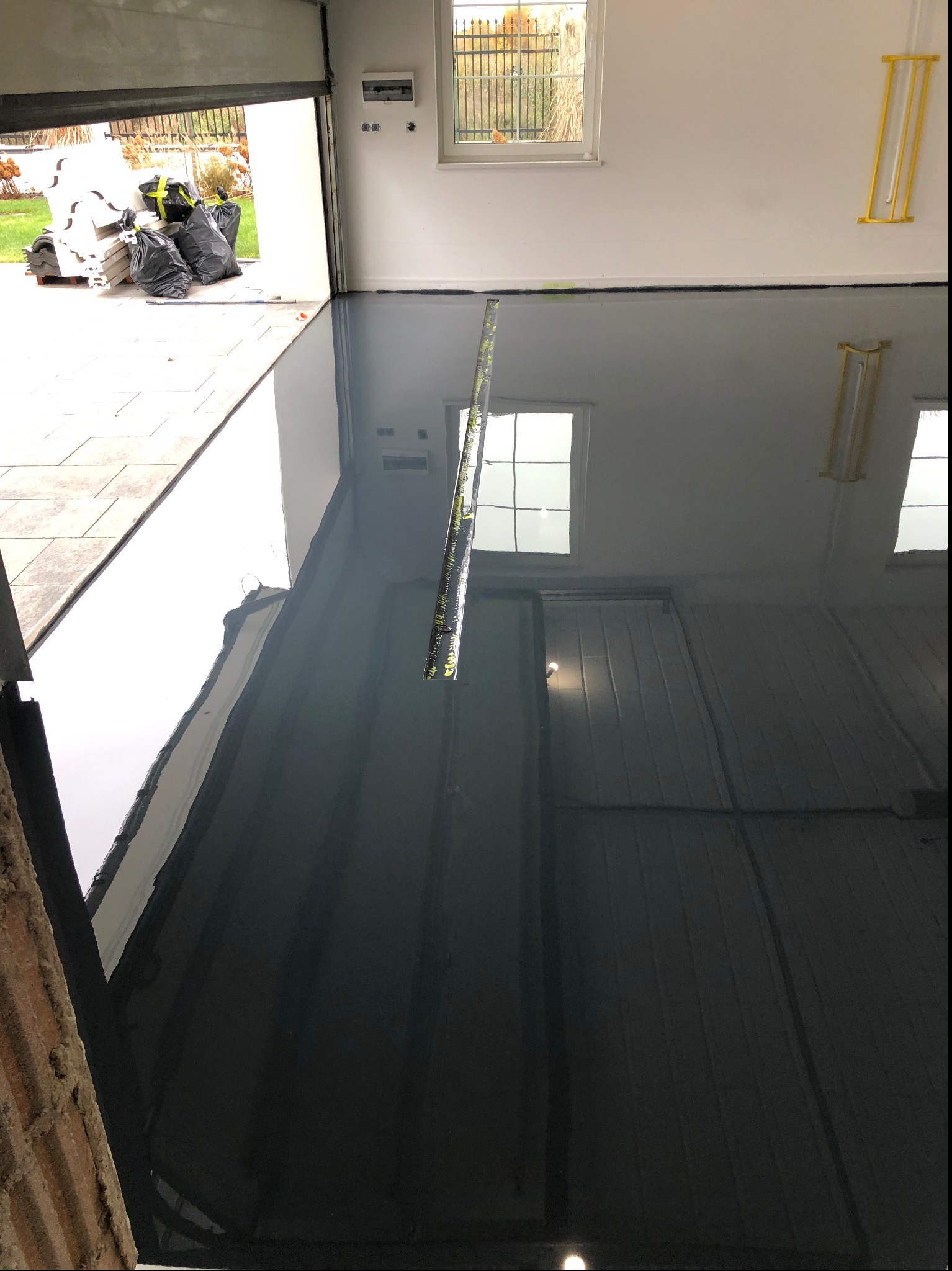 Błyszcząca, ciemnoszara posadzka w garażu z otwartą bramą. Widoczne odbicia okien i lamp. Na zewnątrz trawnik i ogrodzenie. Poziomica leży na podłodze.