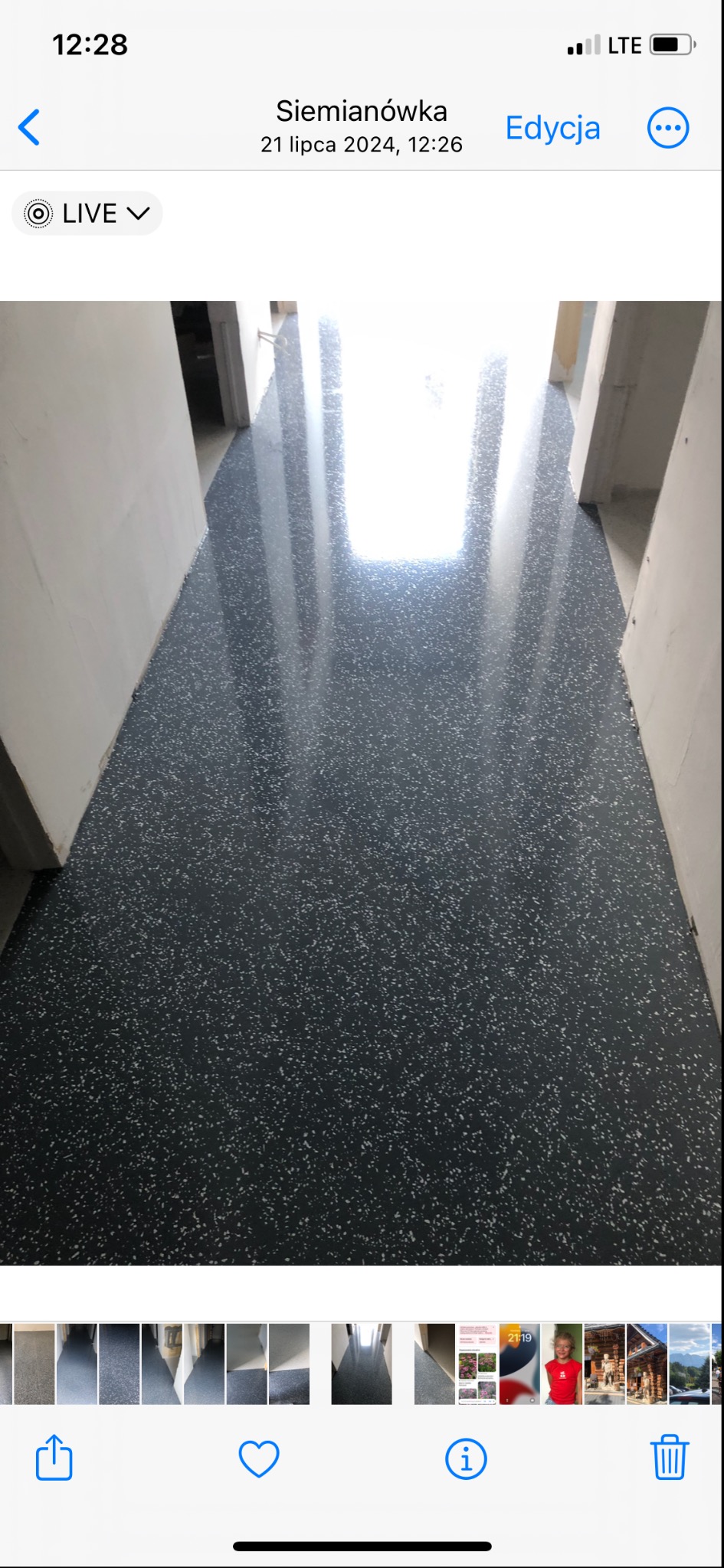 Ciemnoszara posadzka z drobnymi, jasnymi plamkami, wykończona na wysoki połysk. Widoczny fragment korytarza z białymi ścianami i drzwiami. Światło odbija się od powierzchni podłogi.