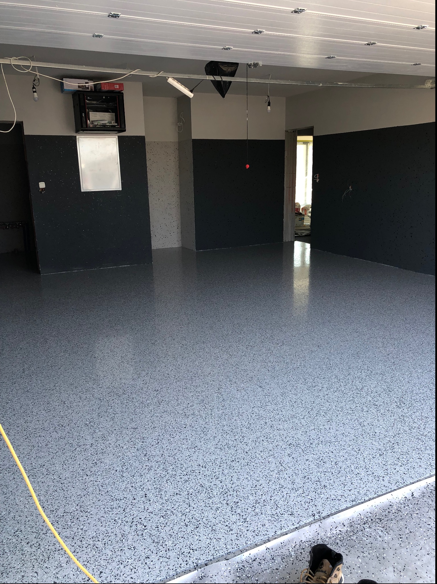 Błyszcząca, szara posadzka z drobnymi, ciemnymi plamkami w garażu. Widoczne fragmenty ścian pomalowanych na szaro i czarno. Żółty kabel na podłodze.