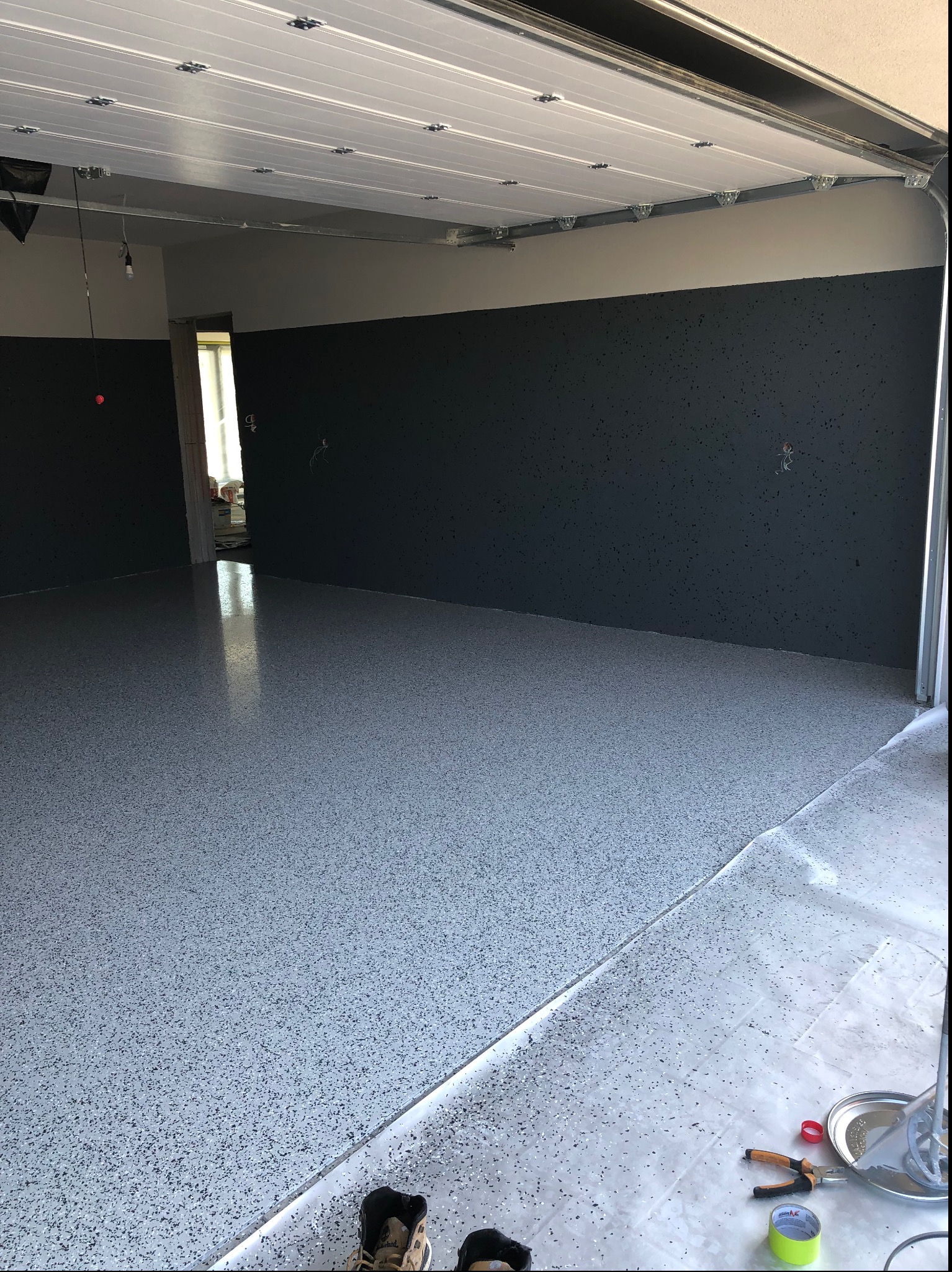Jasnoszara posadzka żywiczna z ciemnymi drobinkami w garażu z otwartą bramą. Widoczne narzędzia i akcesoria do aplikacji żywicy. Ściany pomalowane do połowy na ciemny kolor.