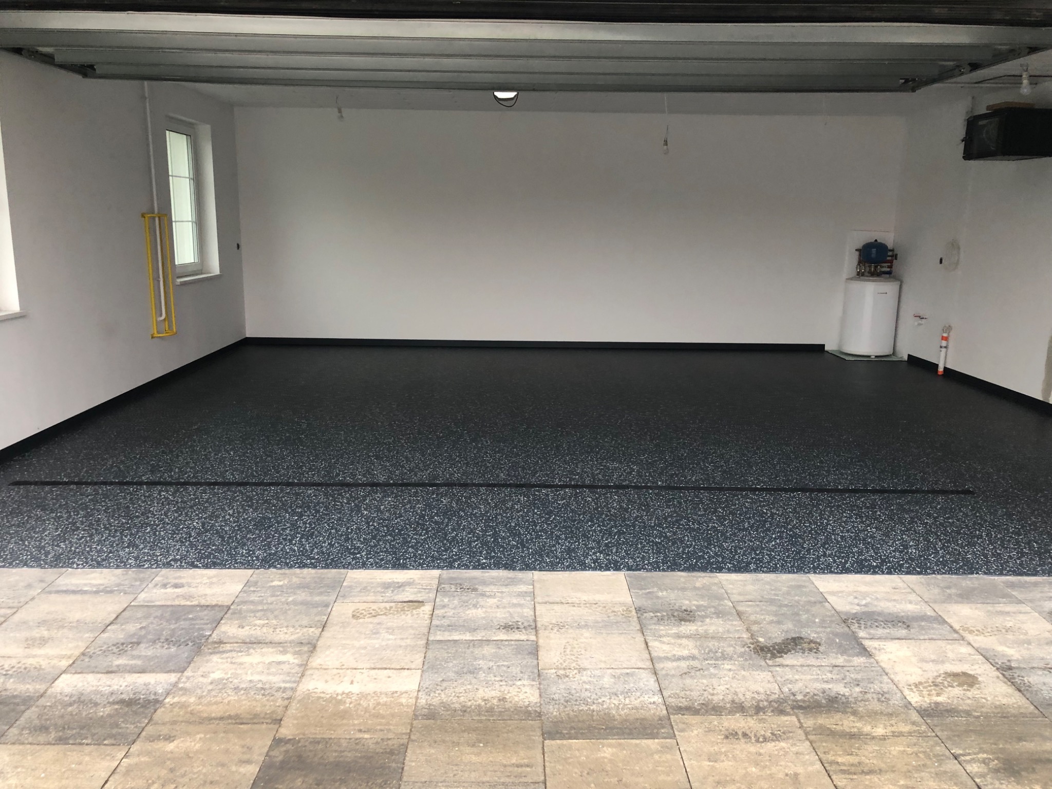 Gładka, ciemna posadzka żywiczna w garażu z odpływem liniowym, widok z zewnątrz na kostkę brukową. Białe ściany, okno i bojler w tle.